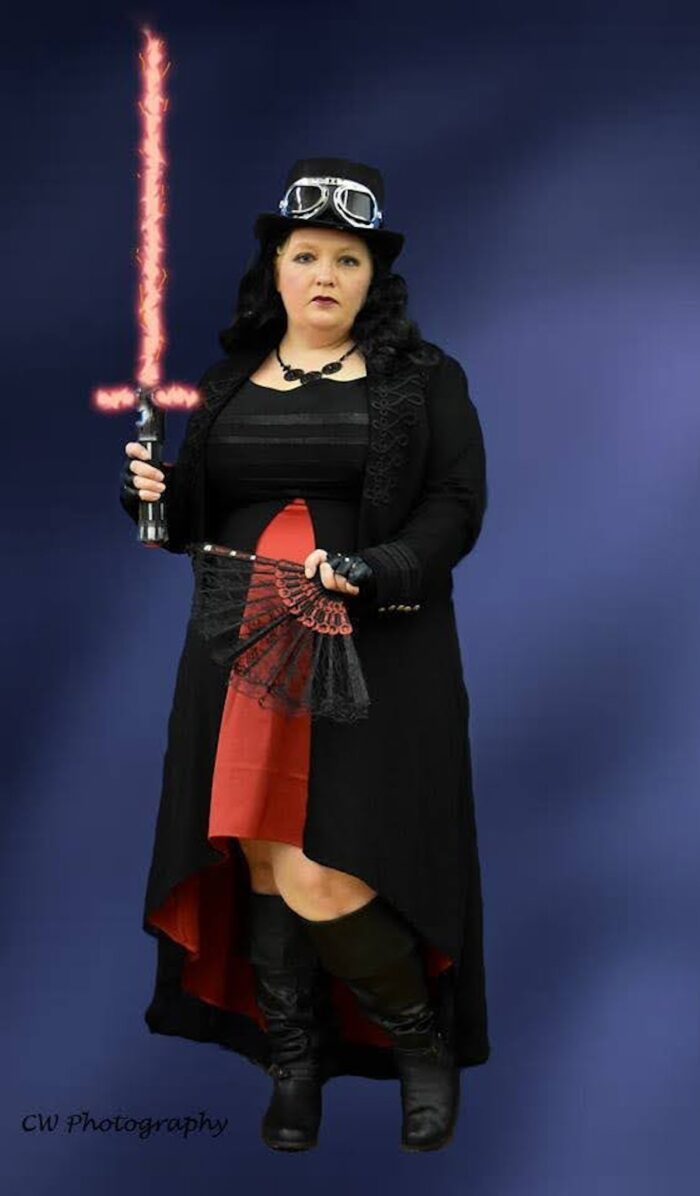 Person holding a fan and lightsaber, wearing boots, Kylo Ren Dress, and Hat with steampunk goggles
